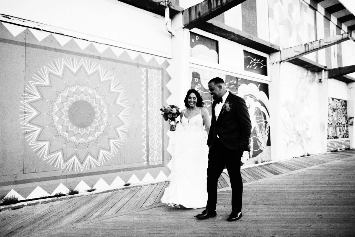 A bride and groom walk together, smiling, along a boardwalk lined with large, patterned murals. Captured by a film photographer NJ style, this black and white image preserves their joyful moment after the wedding ceremony.