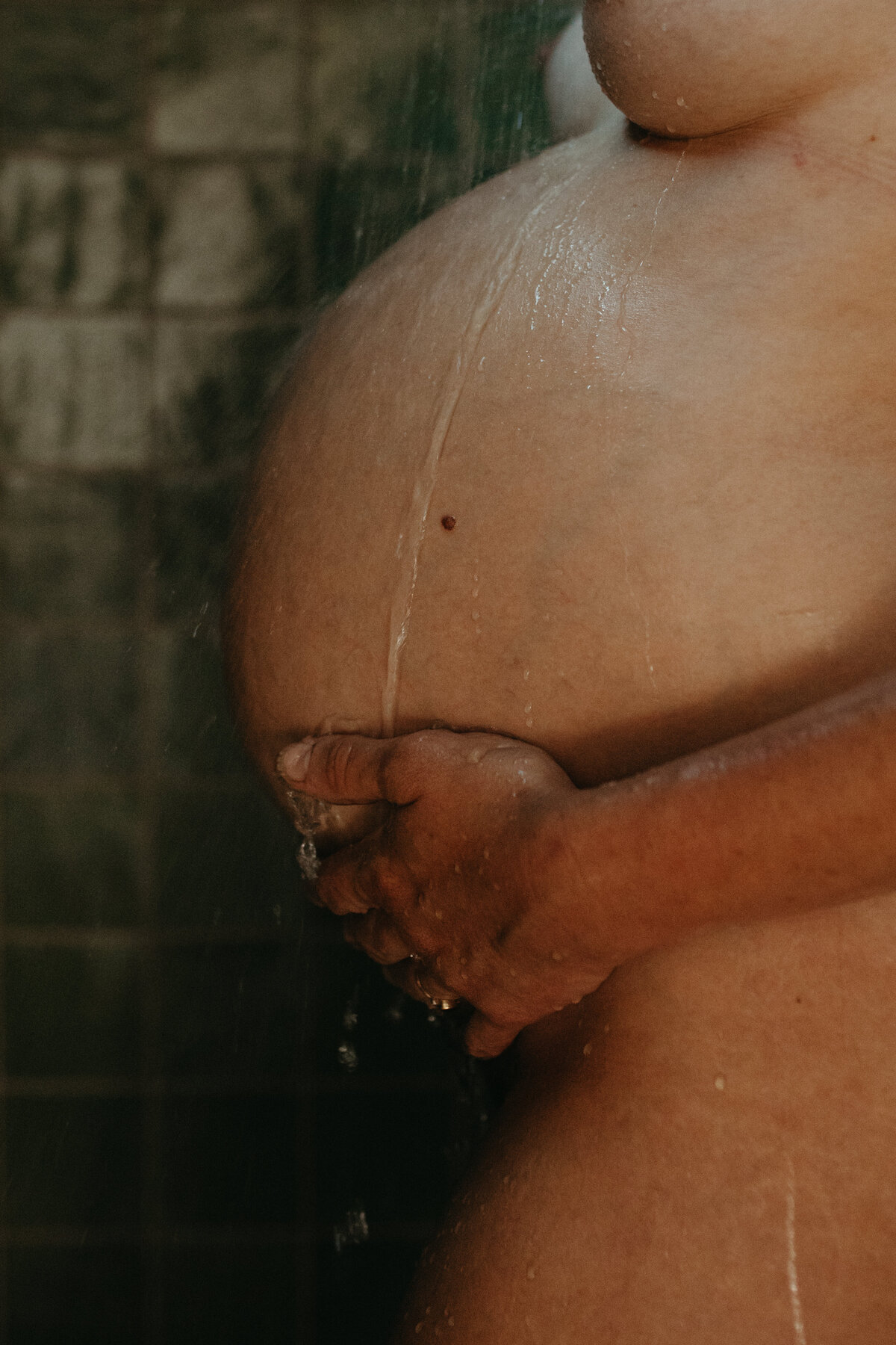 An artistic photo of a pregnant belly in the shower, with water gently cascading over the belly. The natural light and water droplets create a serene and intimate atmosphere.