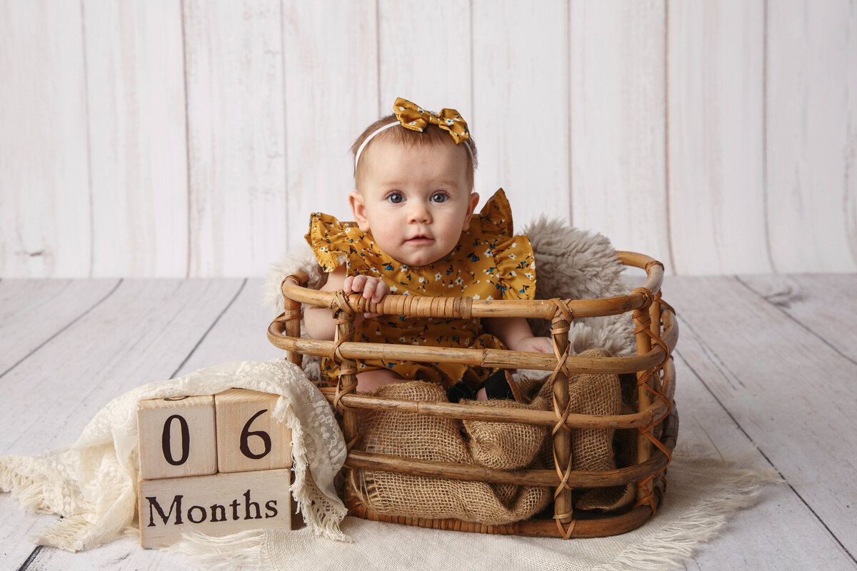 Cute portrait of a six month old baby girl wearing a yellow dress and sitting in a wooden basket photographed by Madison Milestone photographer, Life in Pink.