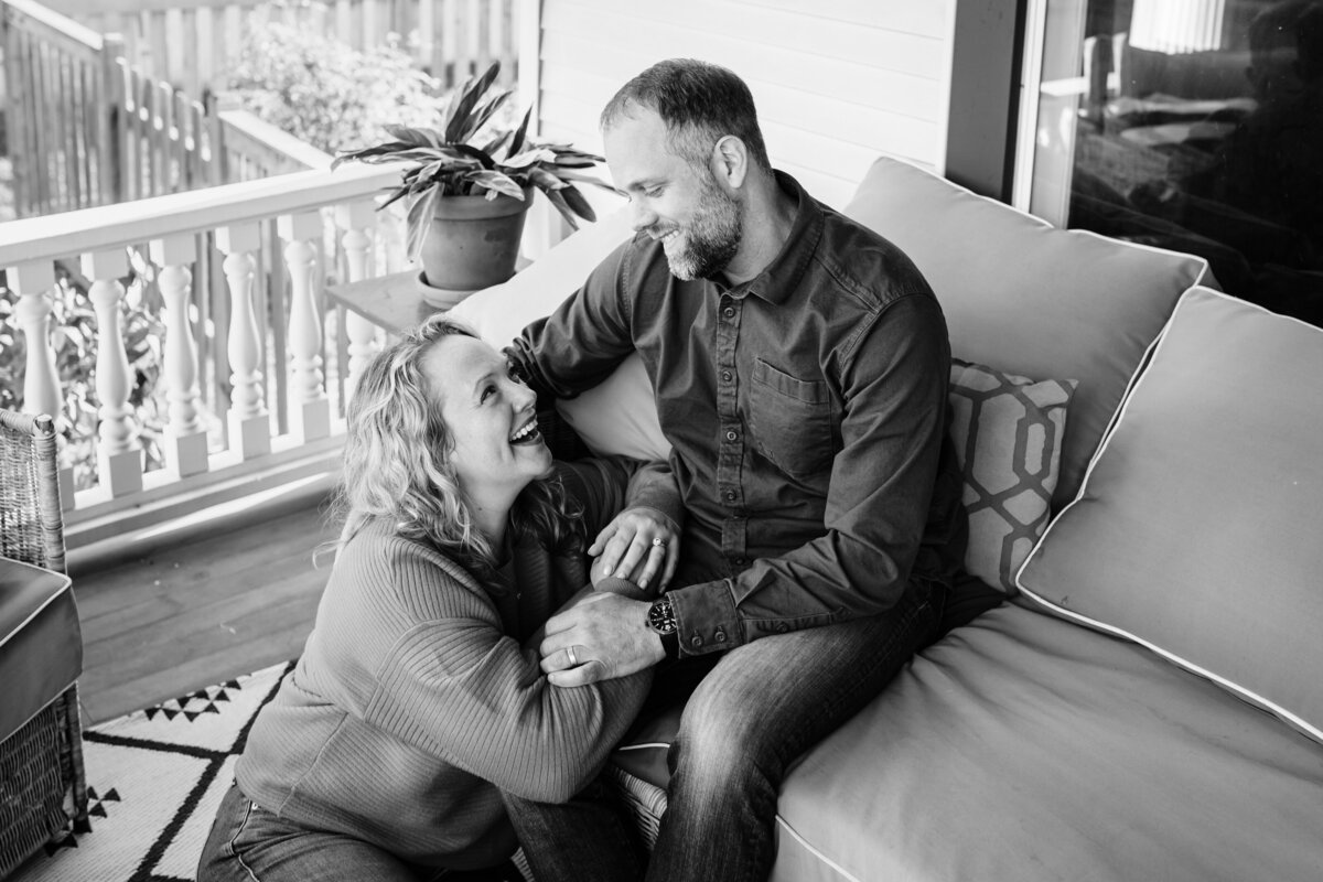 A black and white photo of a couple sitting on a porch, sharing a loving gaze and smile. The woman rests her hand on the man’s arm, both appearing happy and content.
