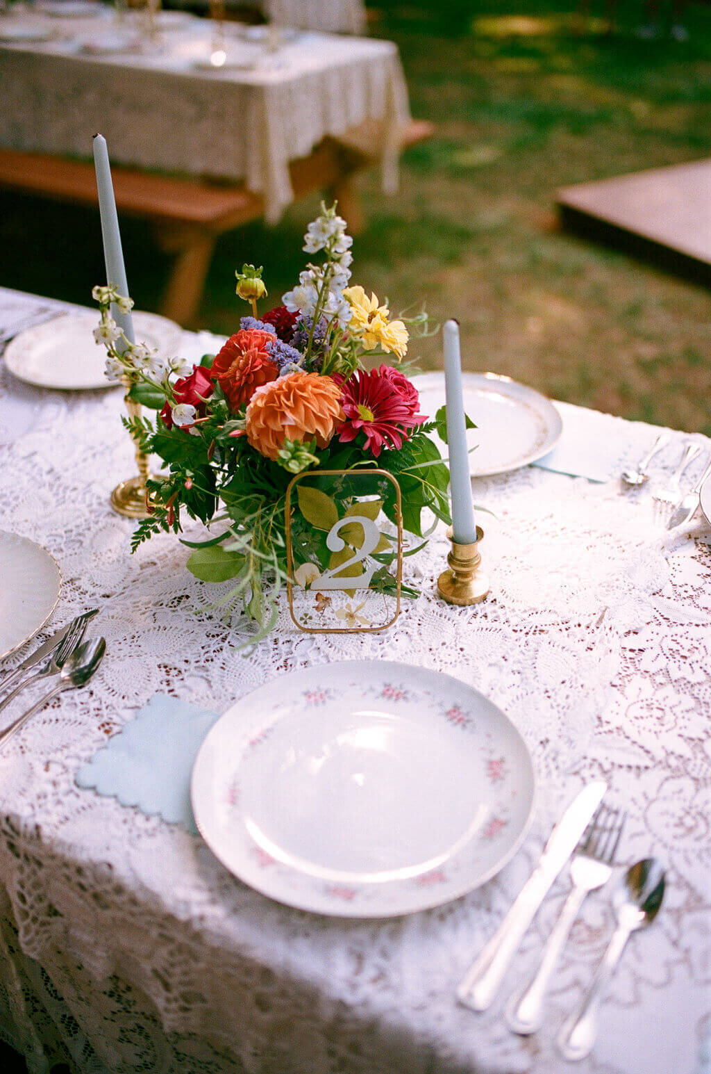 A table setting with lace table cloth, tableware, candles, and flower arrangement.