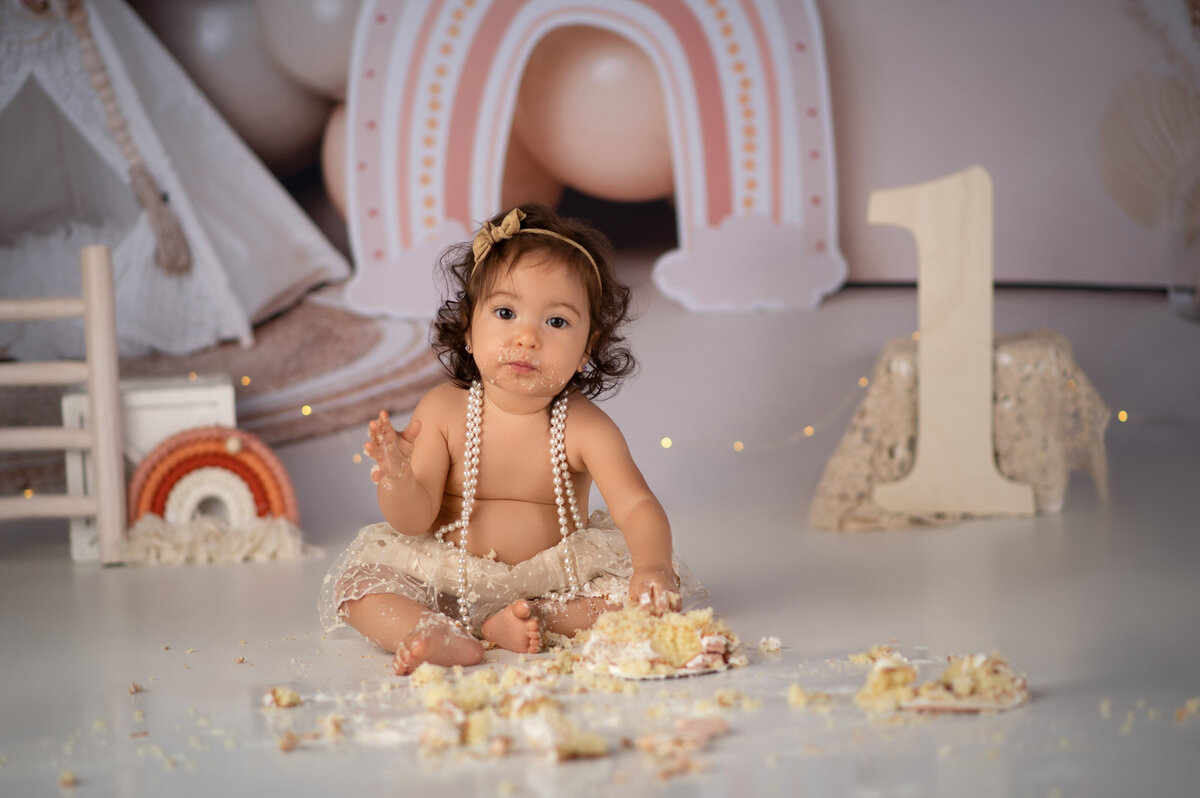 One year old baby girl with long black hair wearing tutu and pearls smashing her birthday cake in front of a boho rainbow backdrop