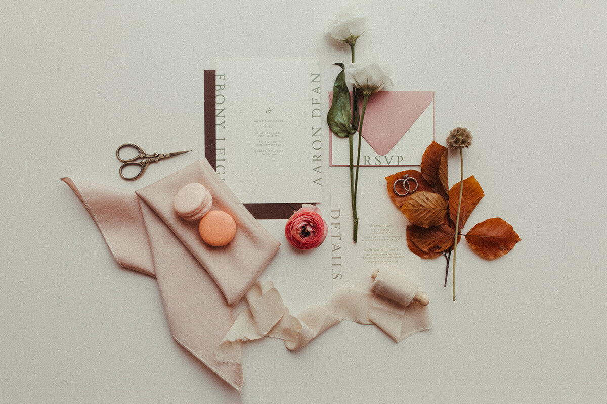 An assortment of white wedding stationery with gray font set with a blush napkin, peach-colored macaroons, and flowers.