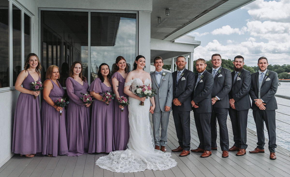 The joyful wedding party, including the bride in her gown and bridesmaids in long lavender dresses alongside groomsmen in gray suits, poses on an outdoor balcony overlooking the water at the Lesner Inn in Virginia Beach, Virginia, captured by Quinn Photography LLC.
