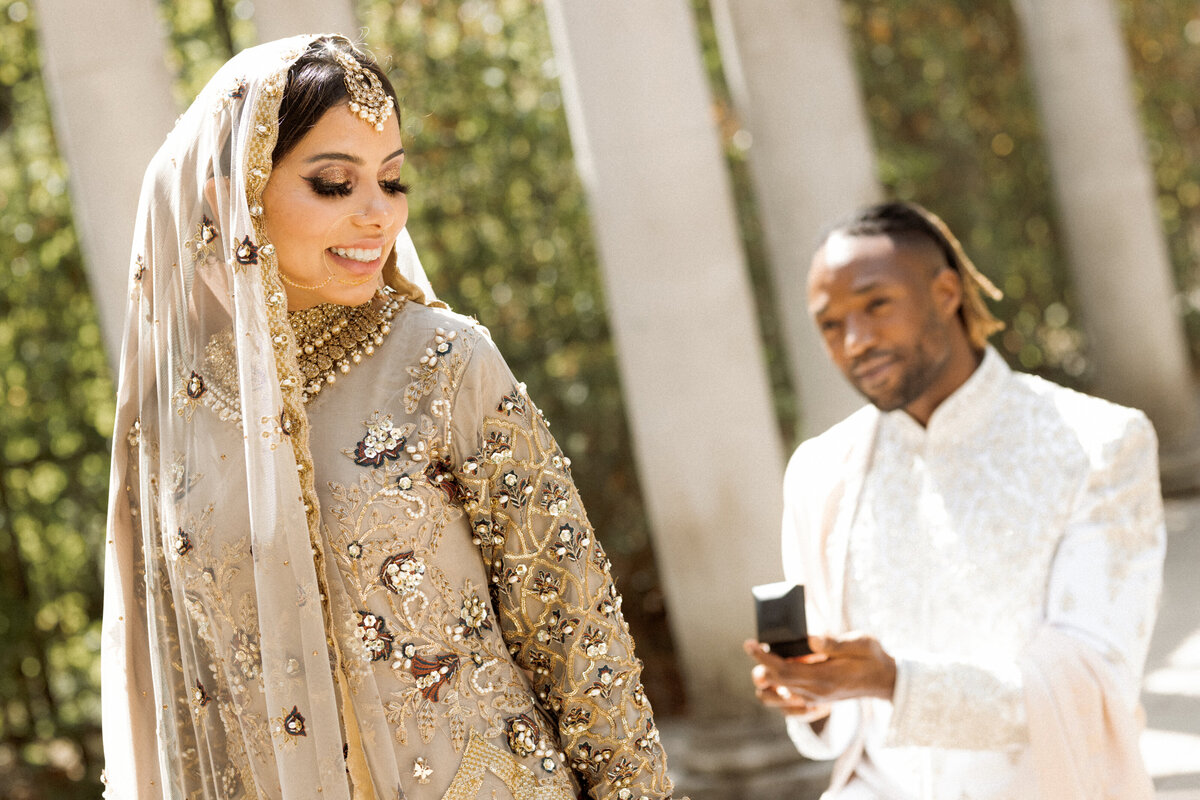 A beautiful lady wearing traditional wedding dress standing with her partner on one knee proposing her at Pergola and hill garden