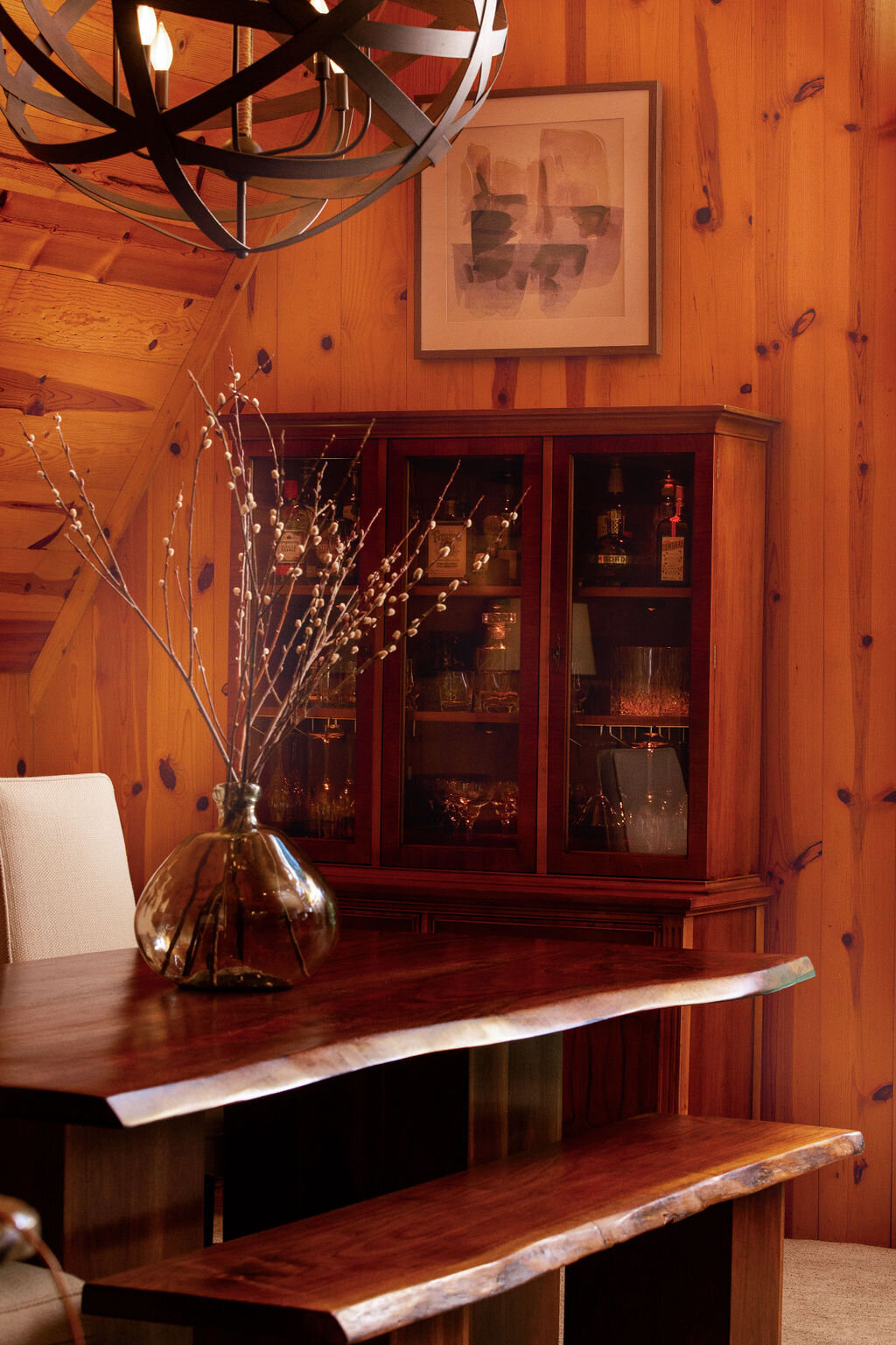 A warmly lit dining area in a wood cabin. A rustic wooden table with a live edge is paired with a wooden bench and chairs with light-colored upholstery. A large, dark glass vase filled with bare branches sits on the table. A dark wood china cabinet with glass doors displays glassware and bottles against the wood-paneled wall. A modern black chandelier hangs above the table, and a framed abstract artwork is on the wall above the china cabinet.