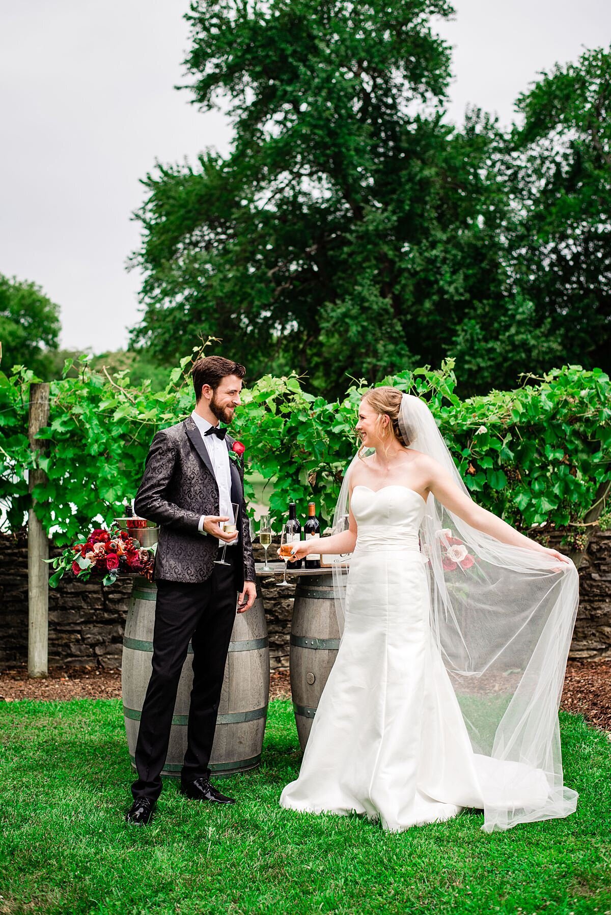 The bride, dressed in a silk strapless wedding dress with a lace bodice, sweetheart neckline and long veil clinks champagne flutes with the groom who is dressed in a black tuxedo with a silk tuxedo jacket embroidered with a paisley pattern and a red rose boutonniere. The bride and groom are drinking Arrington Vineyards Celebration Blanc in front of a barn wood wine bar set on top of two wine barrels decorated with a large bouquet of red roses, blush roses, red anthurium, red anemones, red and pink protea, blue thistle, hypericum berries, dianthus and assorted greenery at their wedding at Arrington Vineyards.