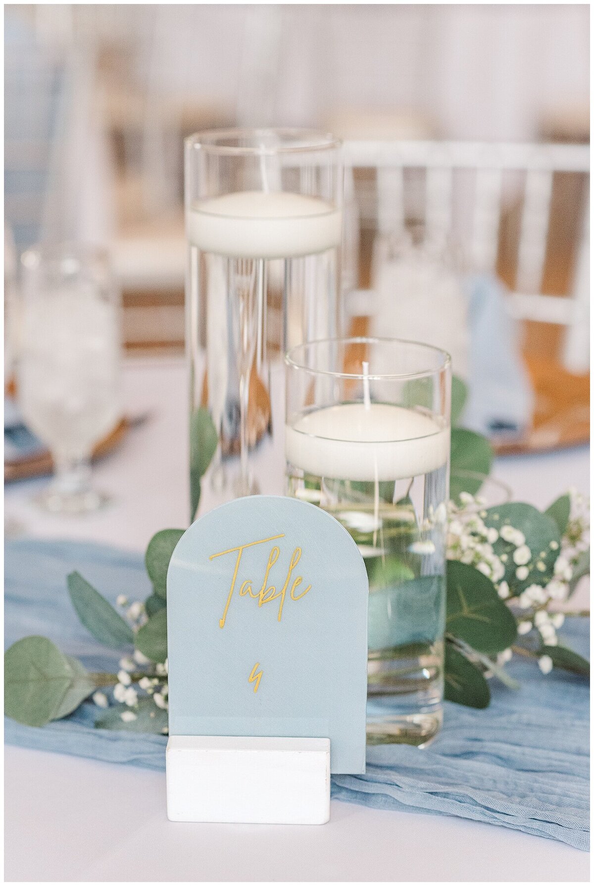 Close up of a reception table with a dusty blue cheese cloth with a dusty blue table sign held up by a white block with 2 different sized candle jars filled with water and eucalyptus and baby's breath flowers surrounding