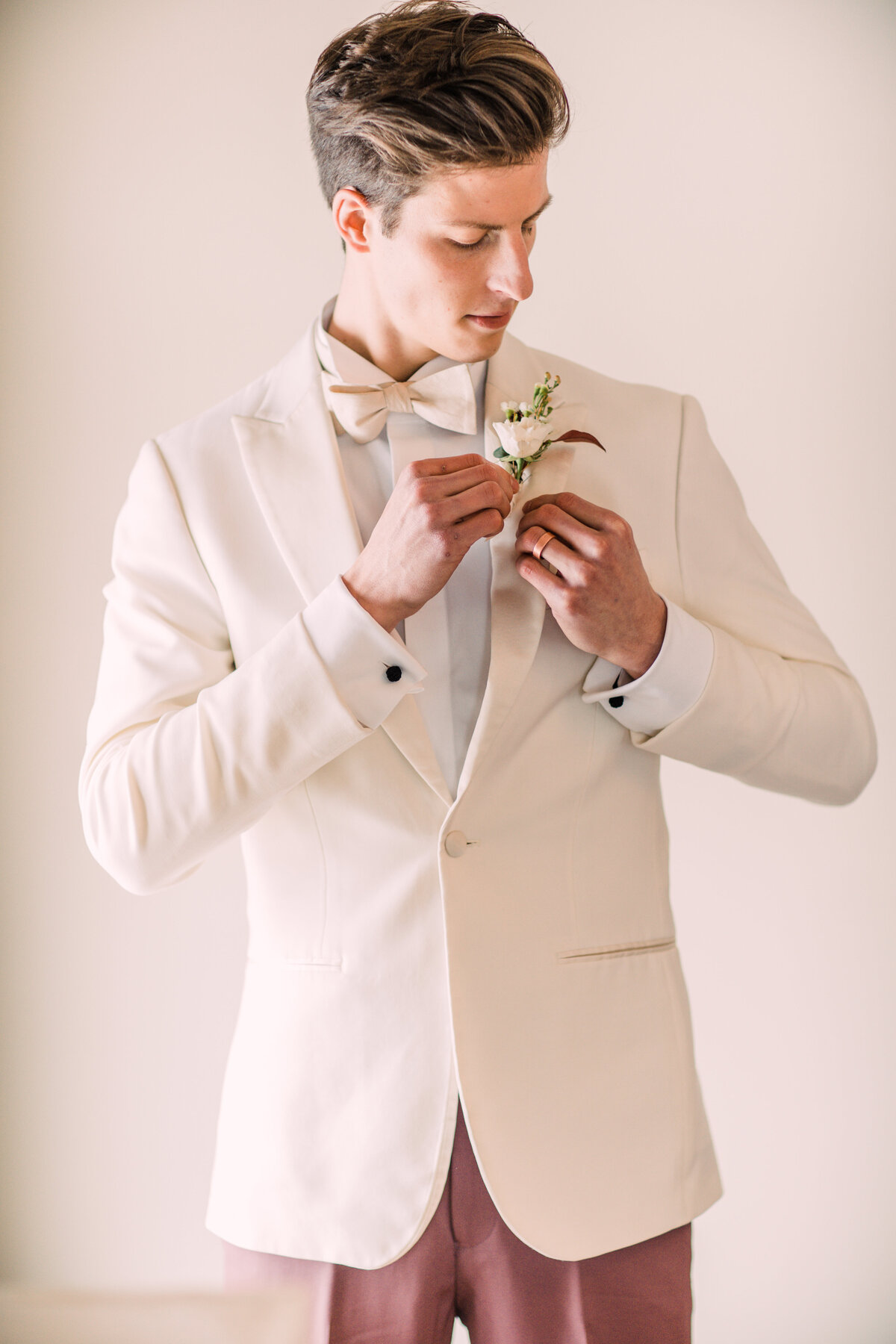 Groom wearing white tuxedo jacket and rose colored pants putting on boutonniere