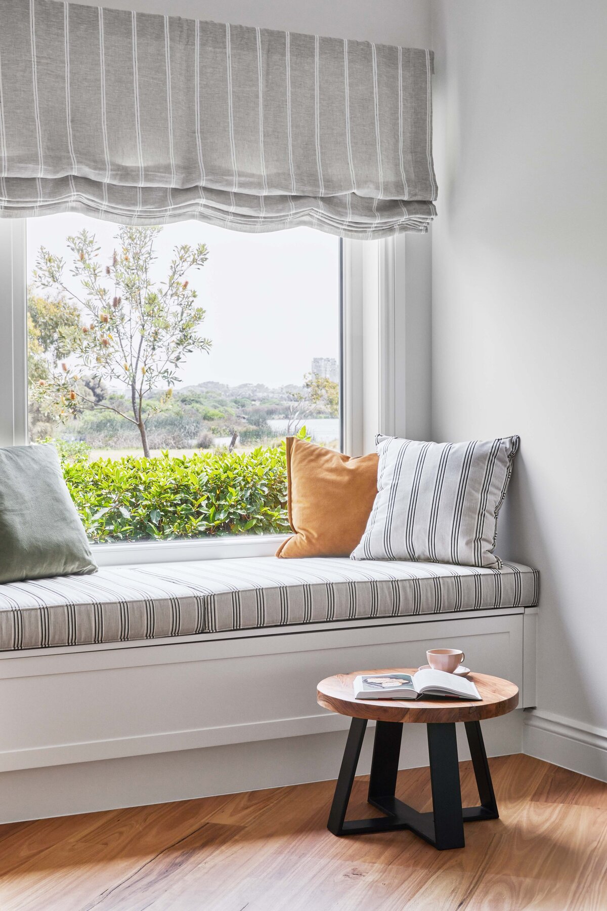 Window seat with striped cushion, soft pillows and garden outlook styled beside a timber side table with an open book and blush mug.