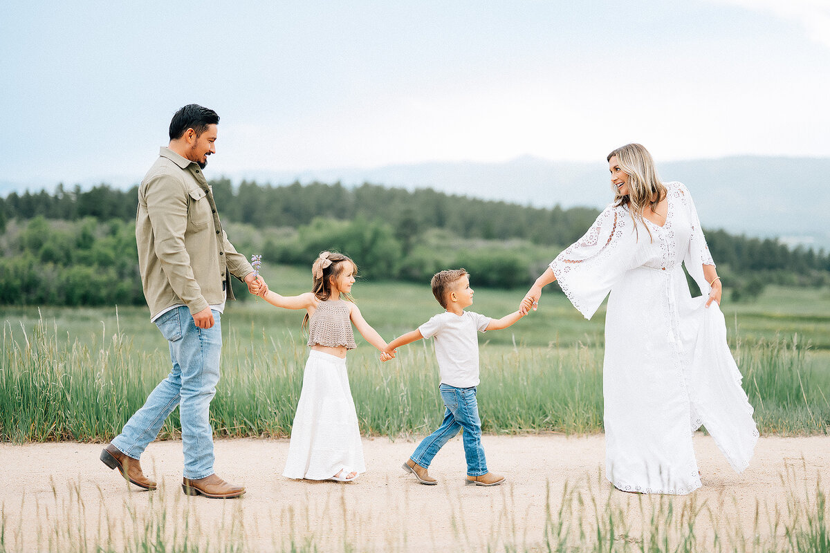 family of four holding hands with mother leading them along the path with mountains in the background 
