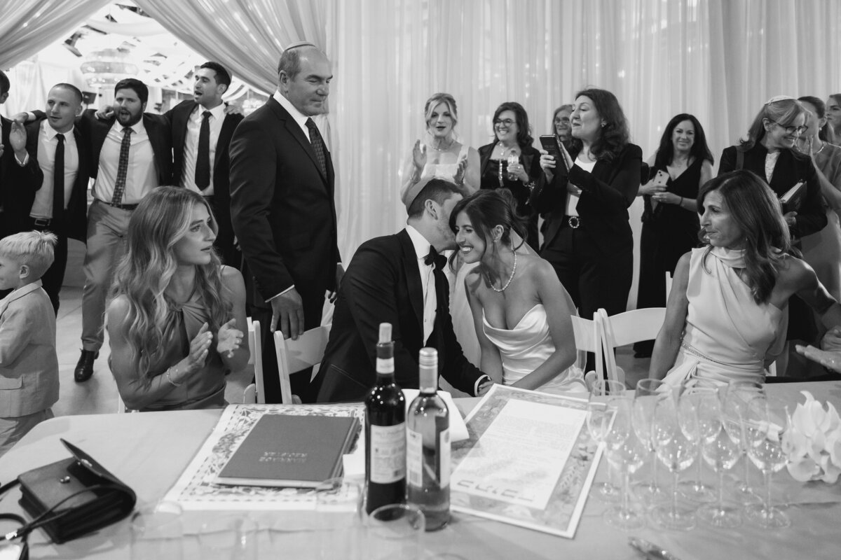 A happy couple signs their ketubah while surrounded by friends and family in black and white and Evergreen Brick Works