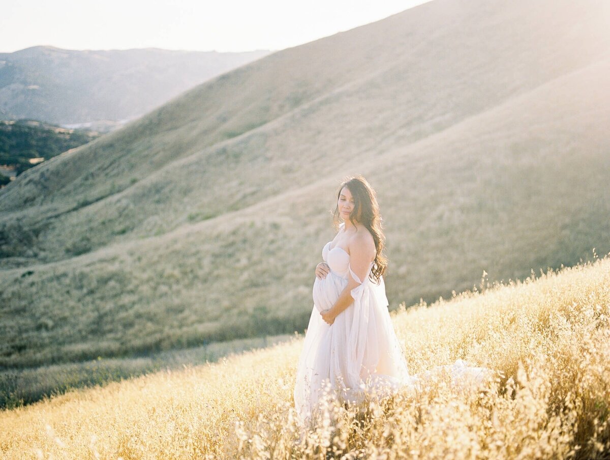 Hillside maternity photoshoot in San Francisco with a pregnant woman standing in a field against hills.