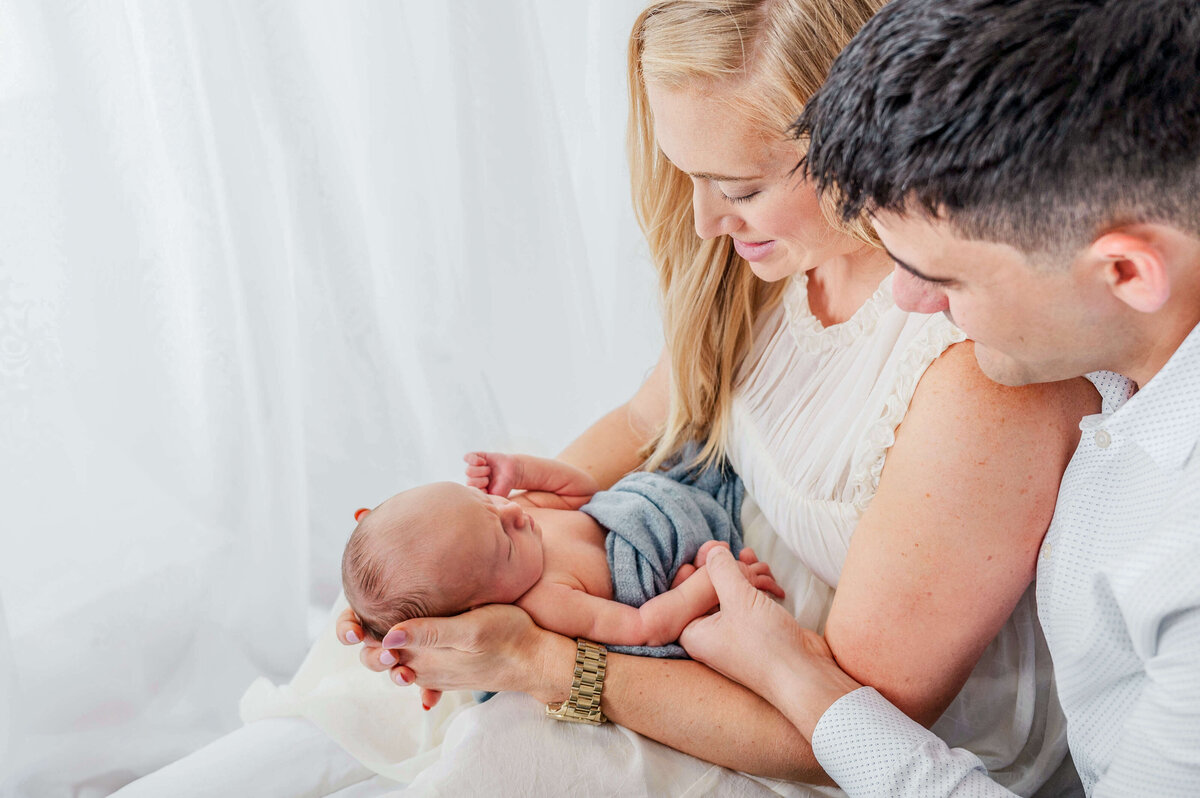 A brunette man looking over his blond haired wife's shoulder at their newborn that she is holding in her arms.