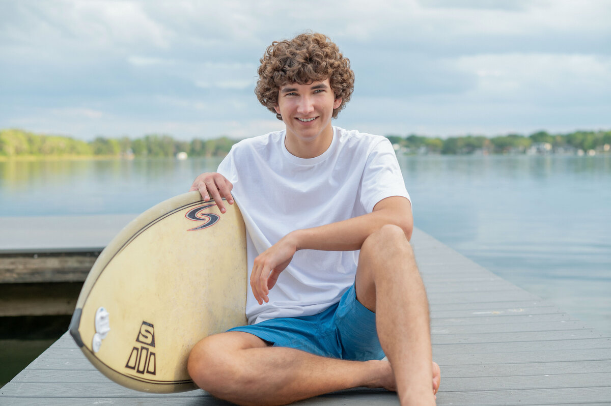 High school senior boy sits on a boardwalk over a lake while holding a surfboard.