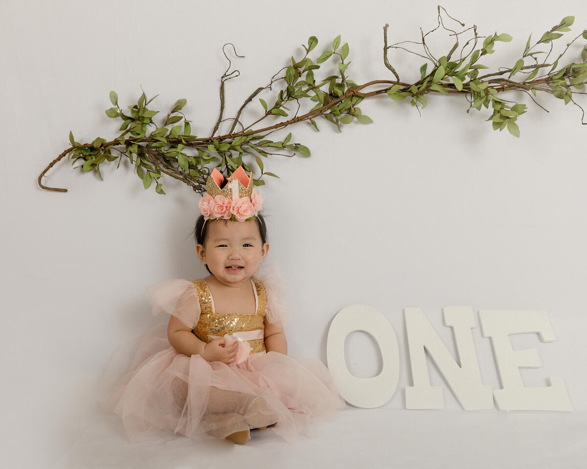 One year old girl with peach dress and crown sitting next to the letters spelling out one.
