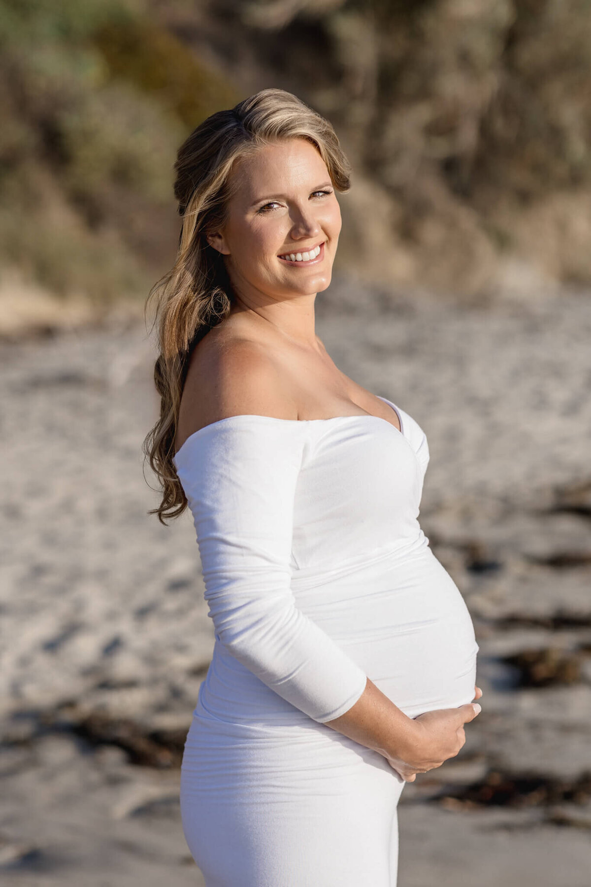 A pregnant woman stands on a beach, smiling, wearing a white off-shoulder dress. The background features sand and blurred greenery.