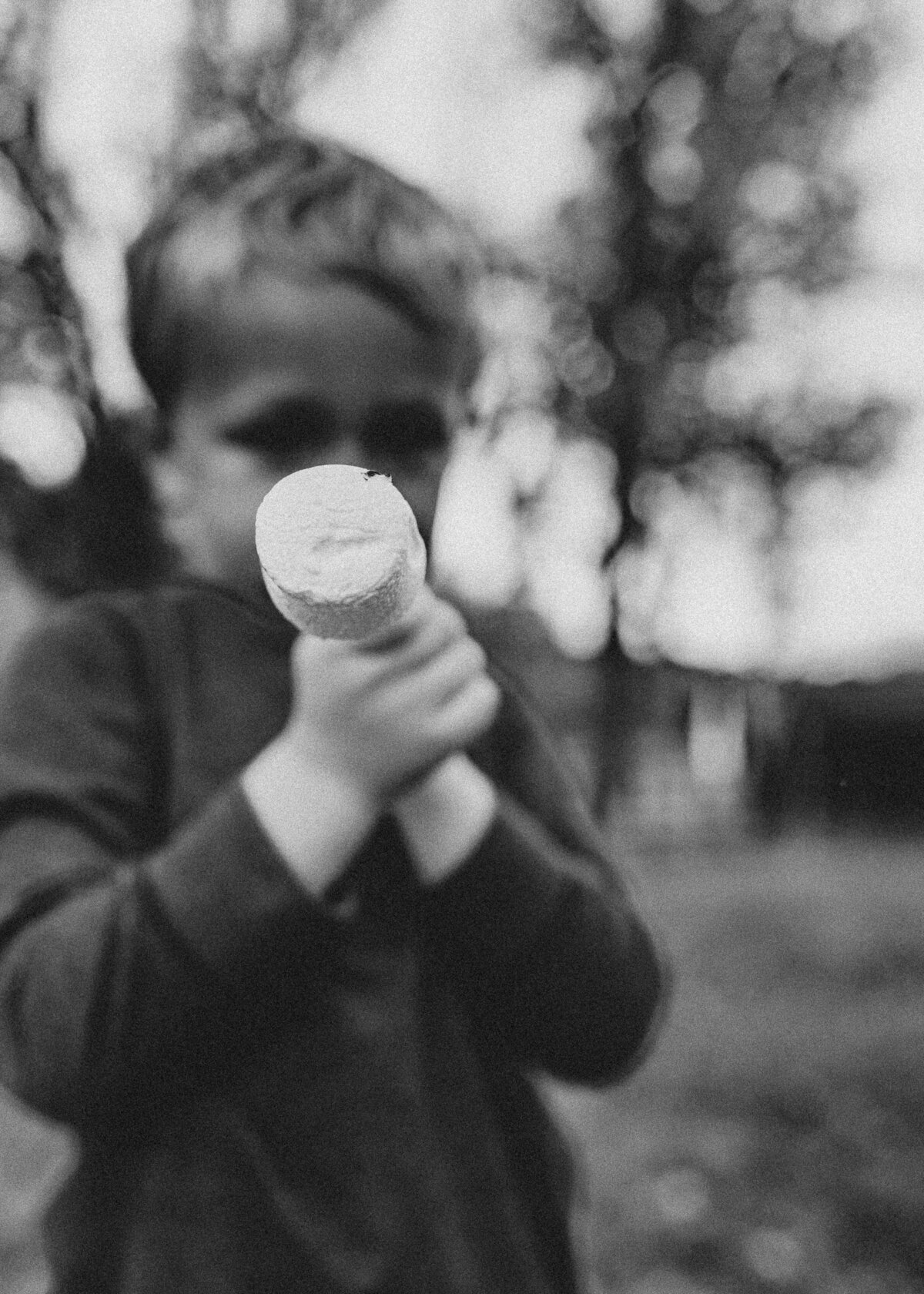 A close-up black-and-white image of a marshmallow on a stick as a young boy shows off what he just roasted.