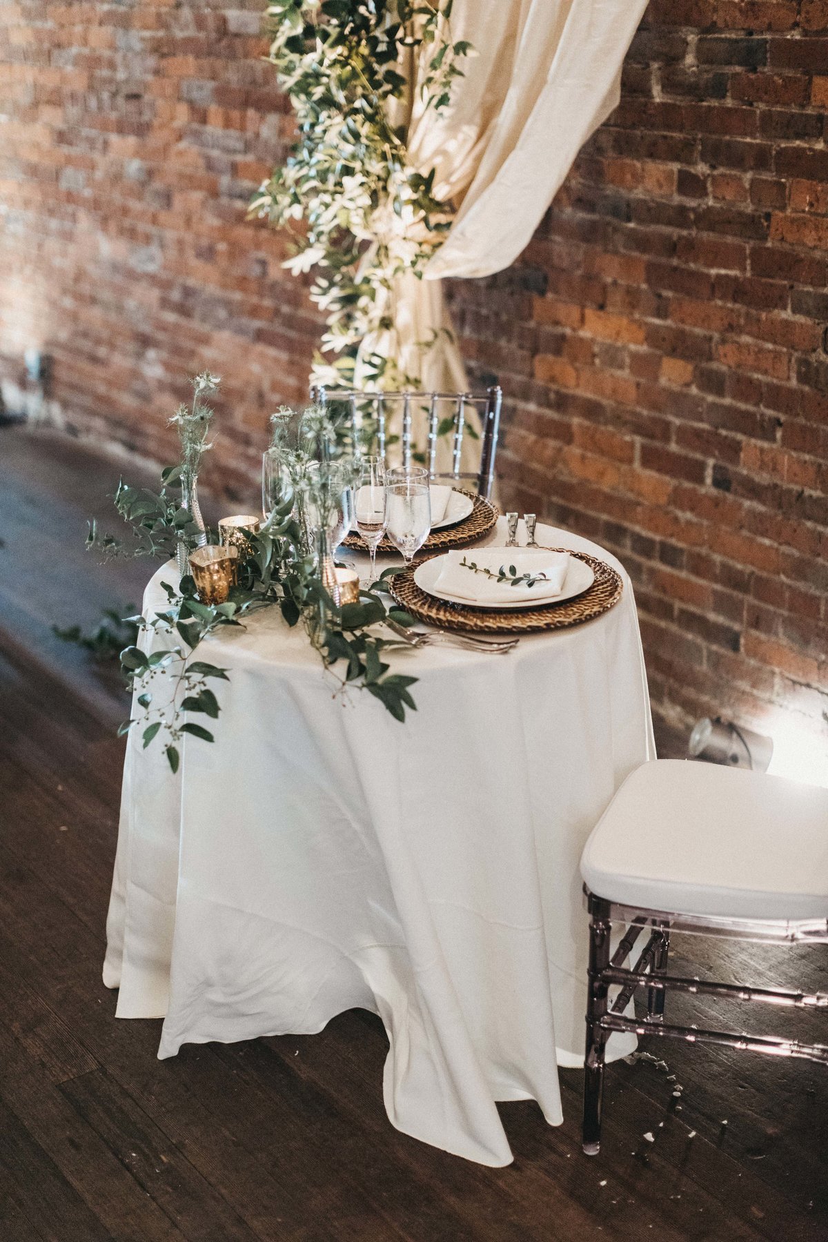 sweetheart table with long white linen, rattan charger plates, greenery centerpiee, and clear Chivari chairs