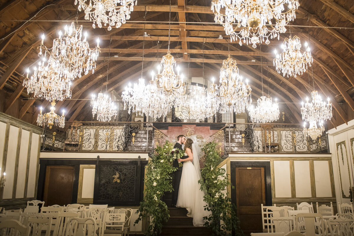 A bride and groom share a kiss in a grand room with chandeliers.