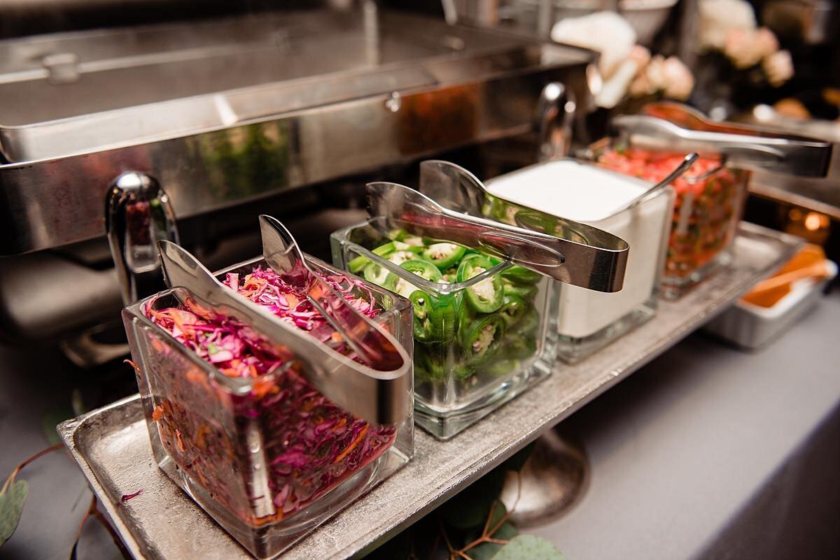 Square glass bowls hold a variety of taco toppings with silver tongs at a wedding taco bar.