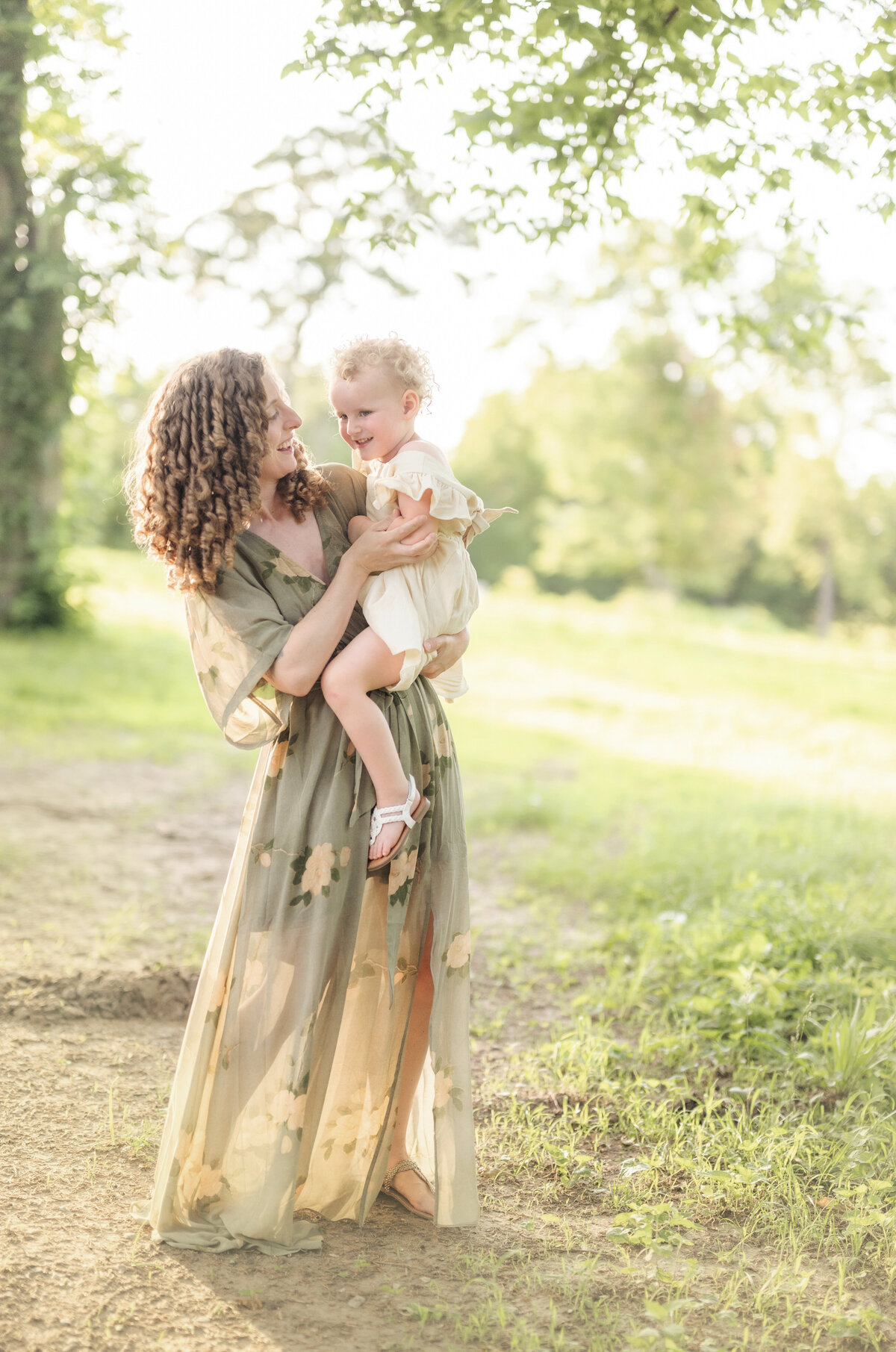 Mother holding her daughter in a long dress while walking along a sun-drenched forest trail.
