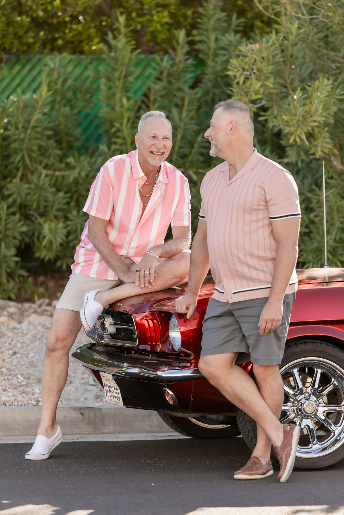 Couple standing together in front of red vintage convertible, framed by lush greenery and golden afternoon light.