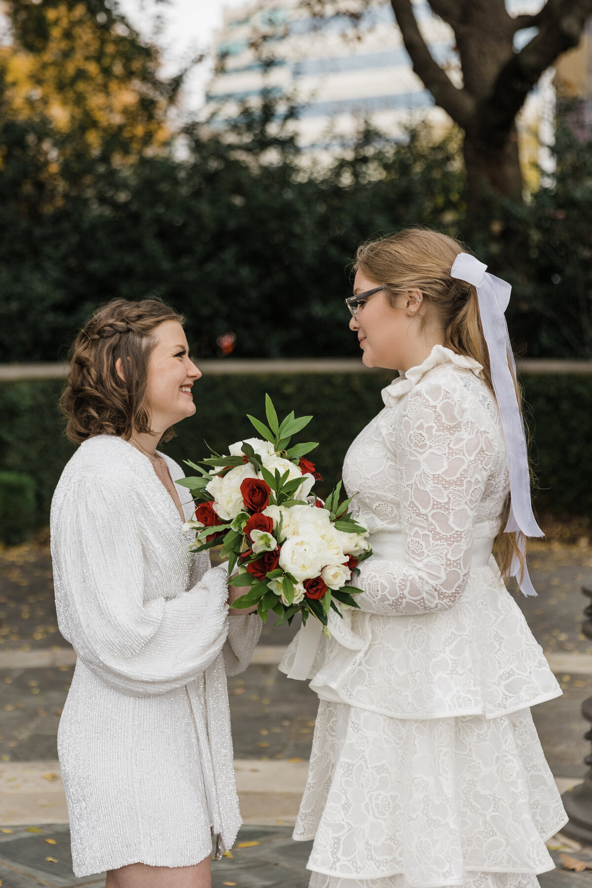A candid shot of two brides sharing a moment before their elopement at Turtle Creek in Dallas, Texas. The bride on the left is wearing a short white dress, and the bride on the right is wearing a long, intricate dress. Both are holding bouquets and smiling at each other.
