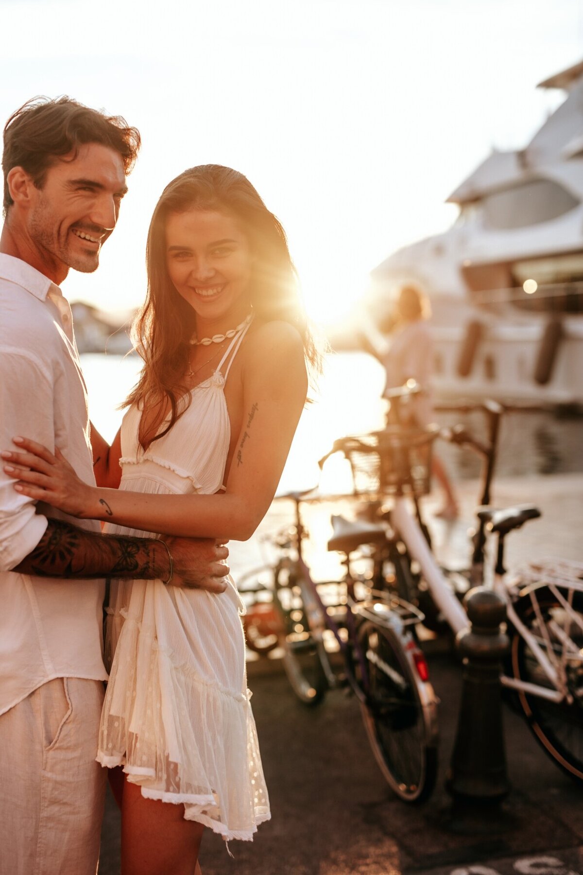 Couple looking at the camera and smiling while embracing each other with the sun coming down behind them and a yacht in the background.