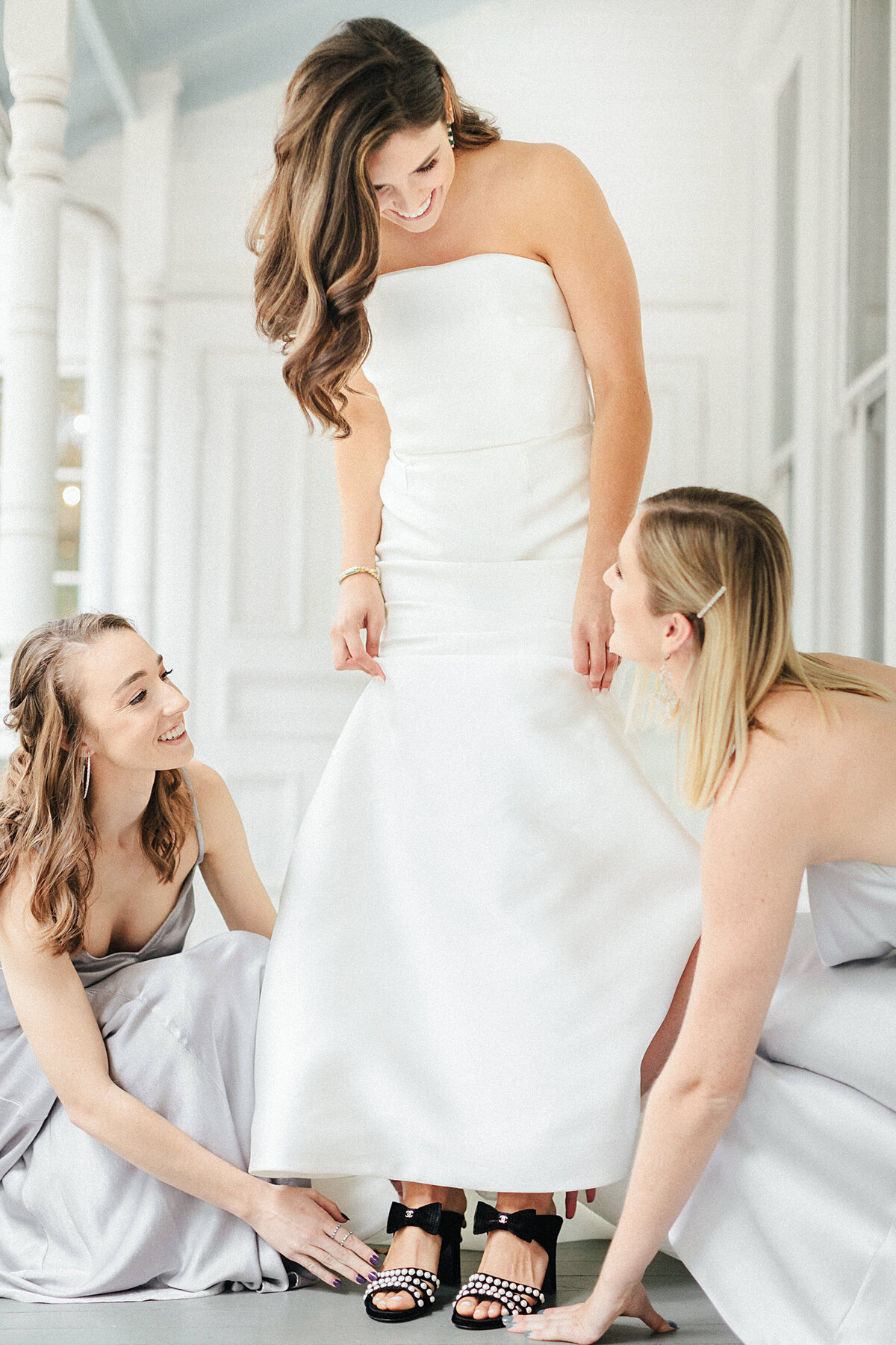 bridesmaids helping bride in white wedding dress put on black heels