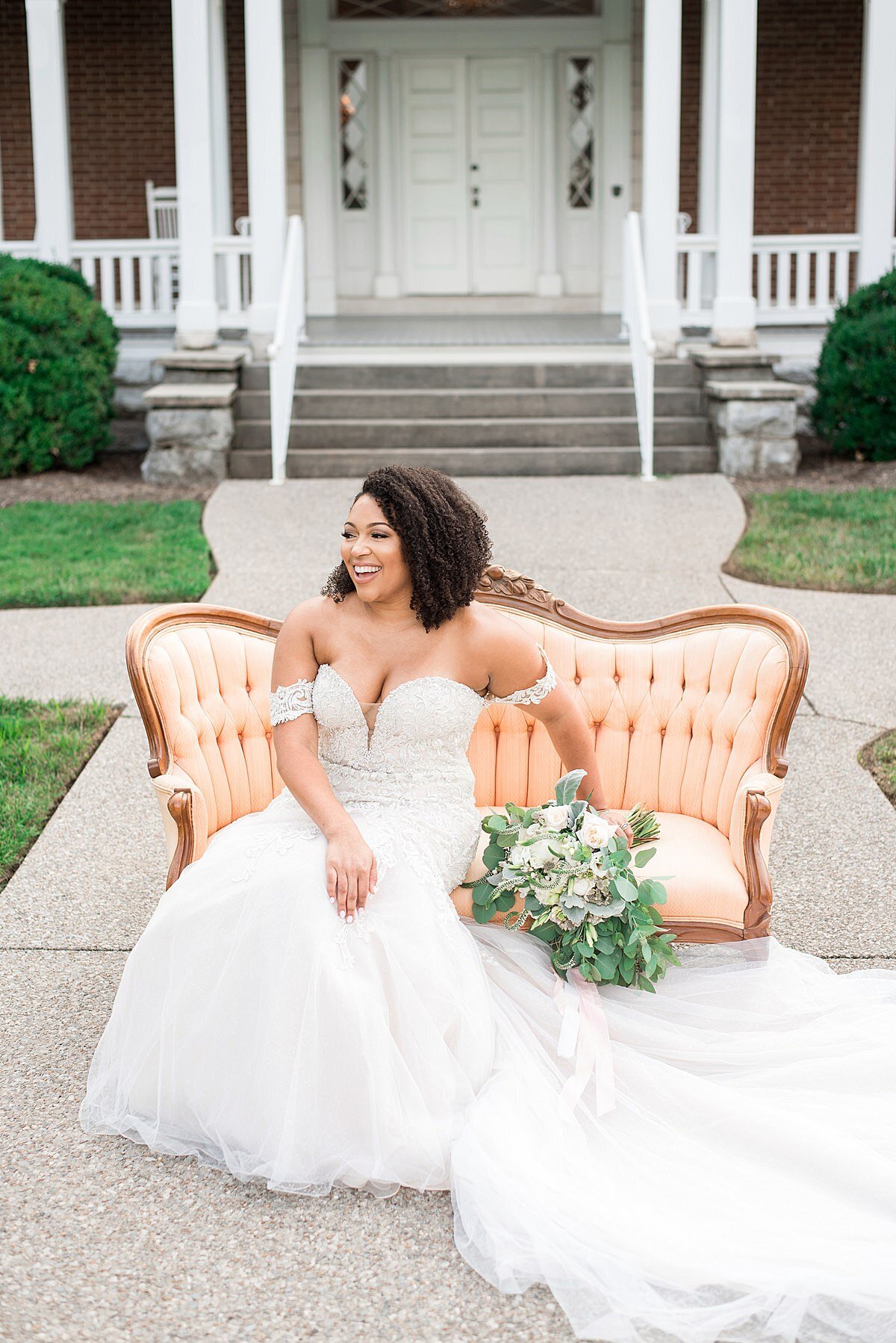 The bride, dressed in an off the shoulder lace mermaid wedding dress with a long train sits on a bright peach silk sofa holding a large bridal bouquet of greenery , white and ivory flowers in front of Ravenswood Mansion