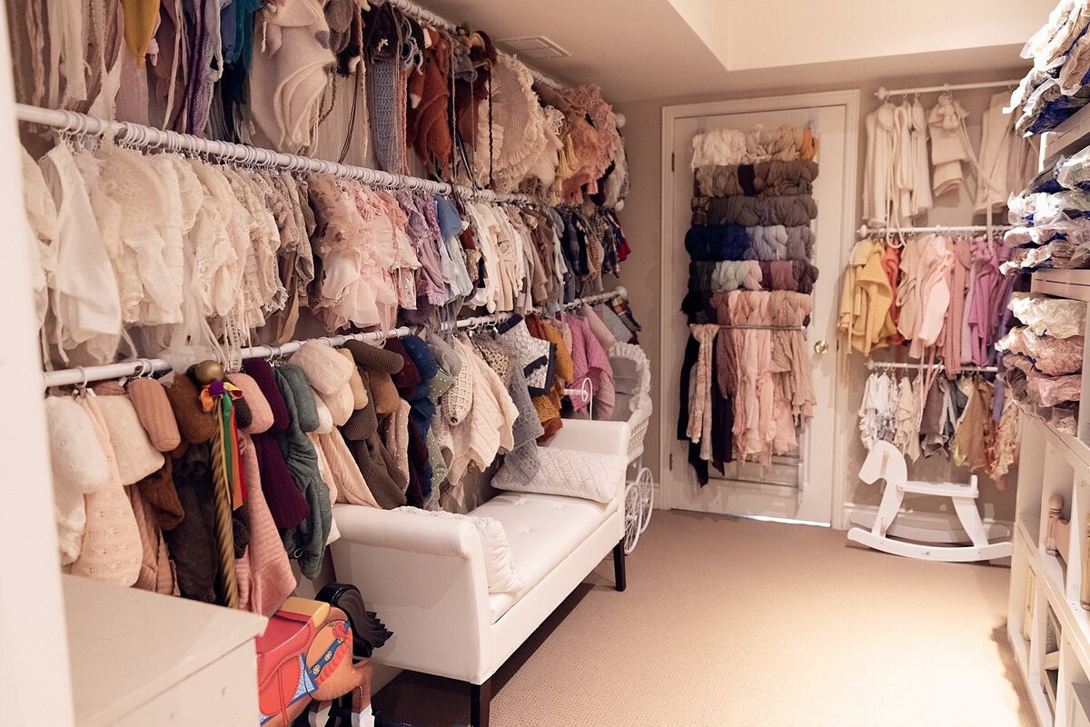 Wide-angle view of a newborn photography studio wardrobe room with shelves of outfits, props, wraps, and an organized seating area.
