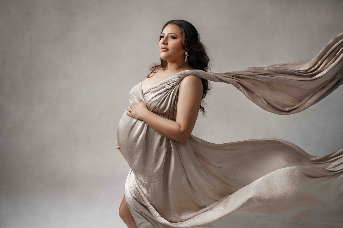 Tacoma pregnant mom stands for maternity photoshoot wearing a flowing silk fabric gown made by photographer Lana Stewart in a travertine stone background. Woman is Ivonne Mendoza of IMArtistry an award-winning wedding professional makeup artist for the PNW area.