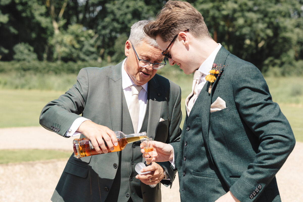 A heartfelt moment captured at Willow Marsh Farm during the wedding day. The groom, dressed in a green suit with a beige tie, shares a laugh with his father, who is also dressed in a matching green suit. The father is pouring a drink from a bottle into a glass held by the groom. Both men are smiling and enjoying a relaxed moment in the warm sunlight, surrounded by the lush greenery of the farm.