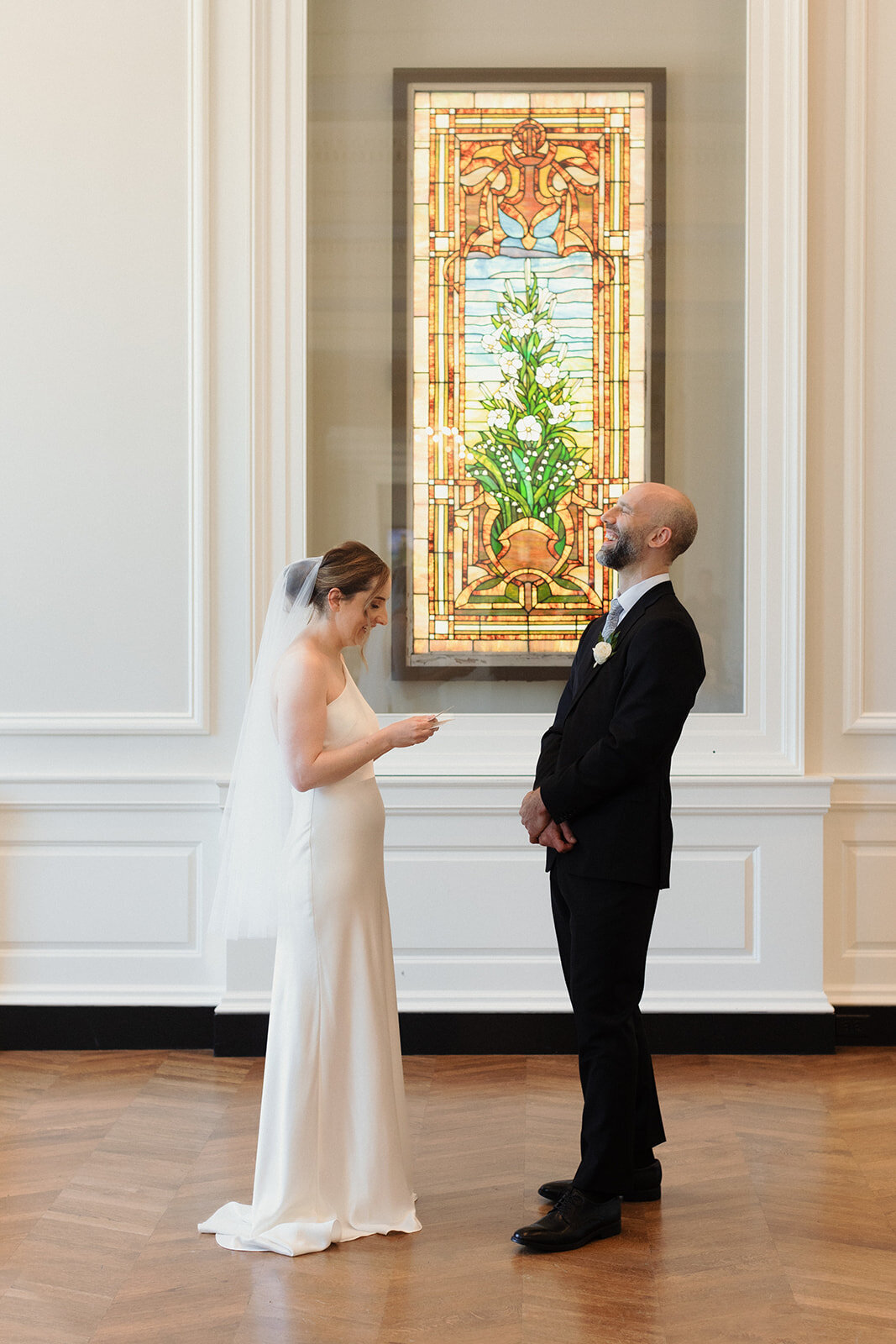 Bride is reading her vows to the groom and he is visibly laughing. Stained glass windows are behind them