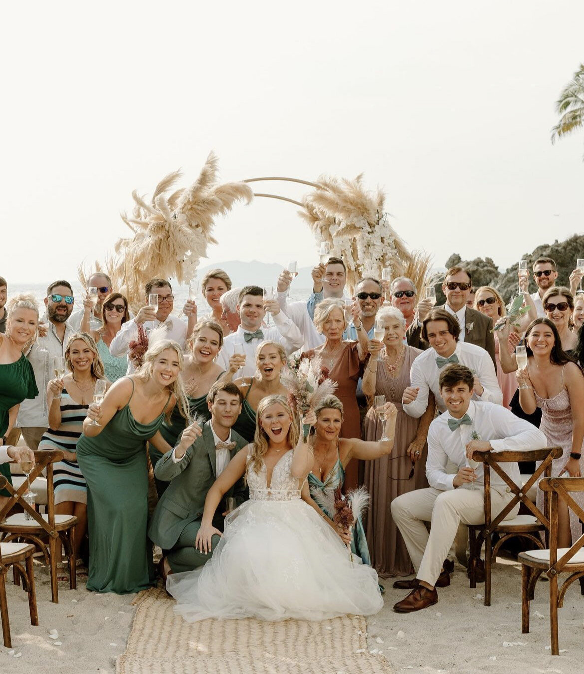 Bride and groom celebrate on the beach with their wedding party and guests, all styled by a mobile bridal hair and makeup team for a flawless destination wedding look.