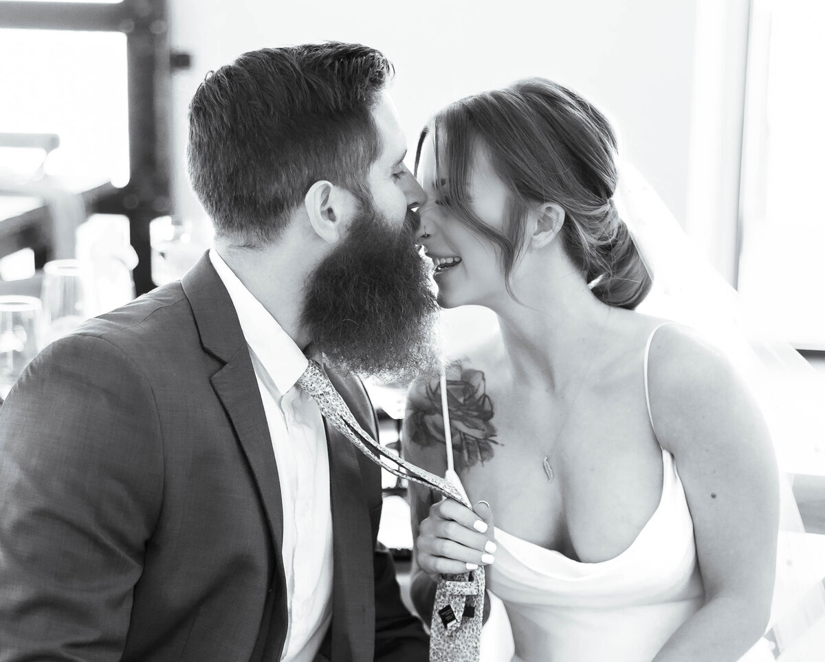Capture the joy and romance of a wedding day with this charming photo of a bride playfully pulling her groom's tie for a sweet kiss, photo in black and white to show the emotion of the moment.
