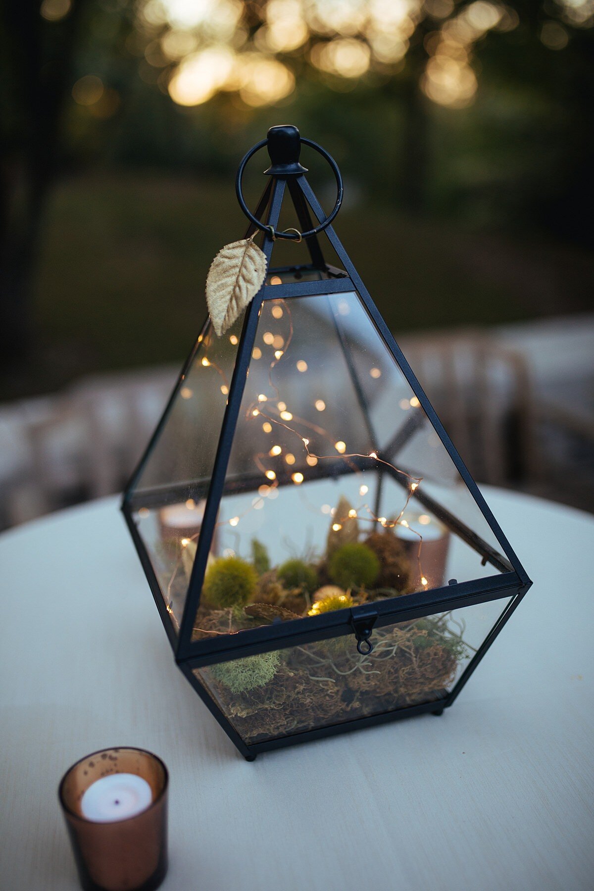 glass terrarium centerpiece for cocktail table filled with moss and fairy lights on a table next to a copper votive candle