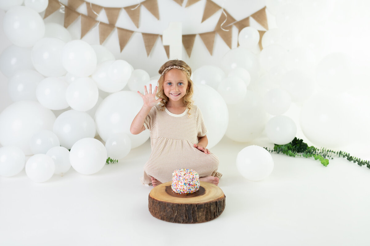 Milestone birthday portrait of a beaming five-year-old waving during her cake smash—celebrating with joy and sprinkles.