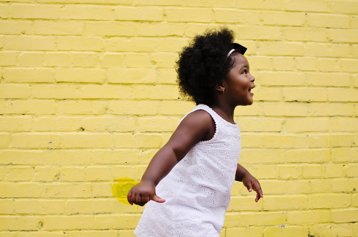 Portrait of a little girl laughing against a yellow brick wall in Old Town Alexandria taken by Sarah Alice Photography