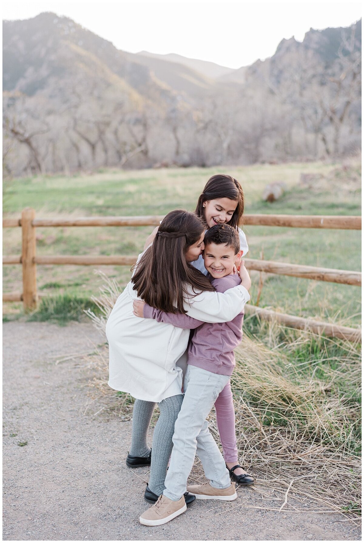 Two young girls in white tops hug their brother in a purple sweater as he smiles and squirms with the boulder flat irons in the background