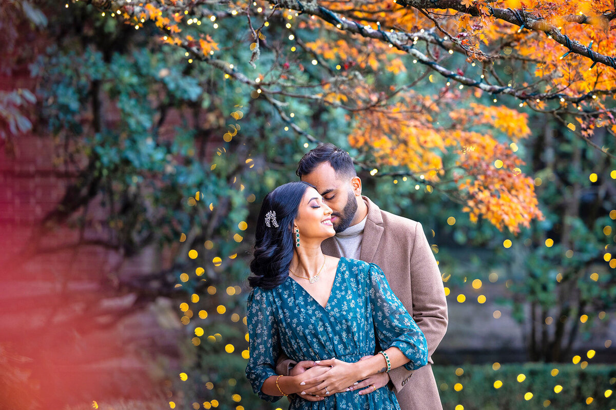 Man holds a woman from behind as they nuzzle their heads together surrounded by Christmas lights and autumn trees in an engagement session at Filoli. Photo by Sacramento wedding photographer, philippe studio pro.