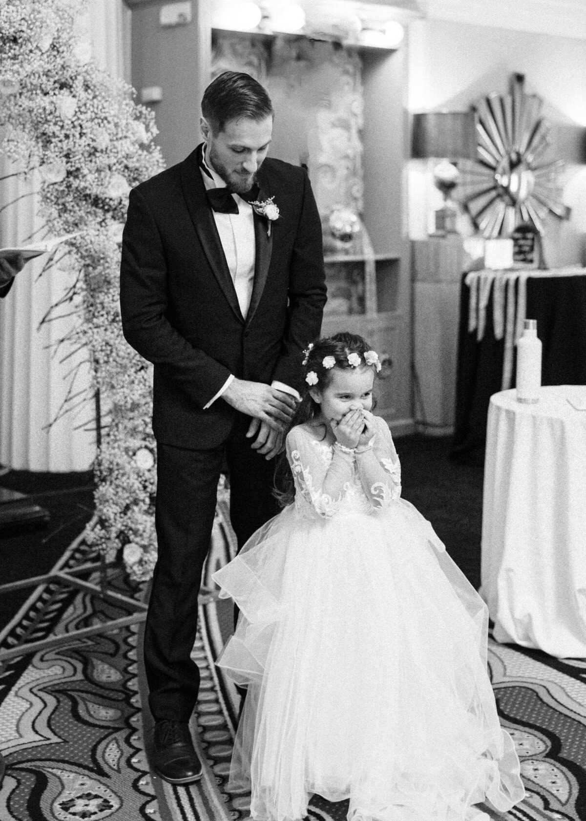 A man in a suit adjusts a young girl's dress at a formal event.