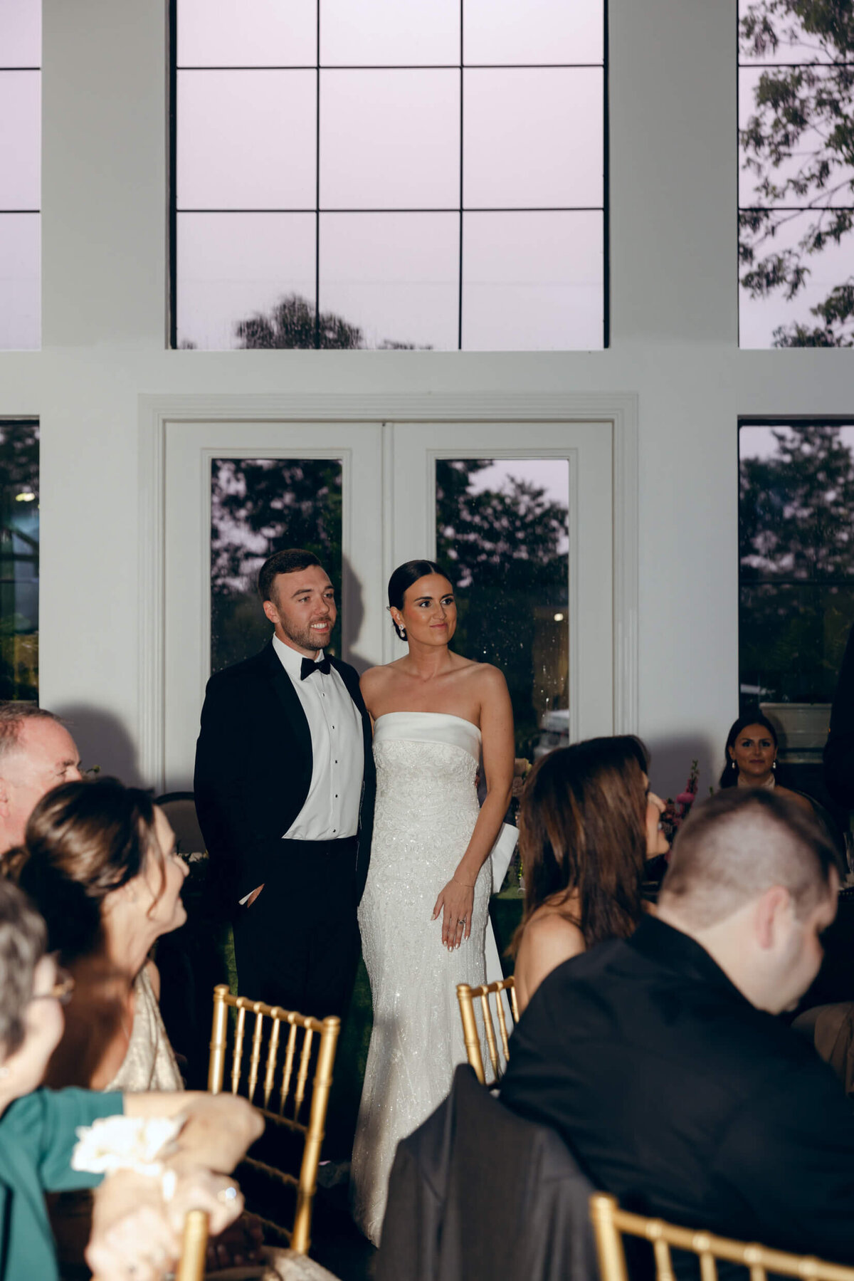 A candid wedding photo fo the bride and groom looking at their friend giving a speech at their wedding reception
