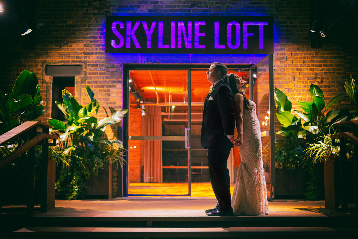 A bride and groom are standing in front of a skyline loft sign.