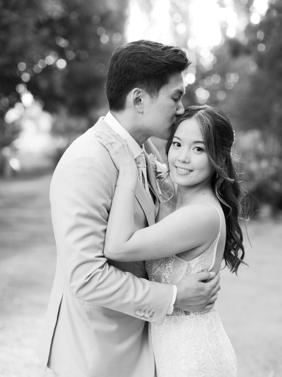 A black-and-white wedding photo of a couple embracing outdoors. The groom kisses the bride's forehead.