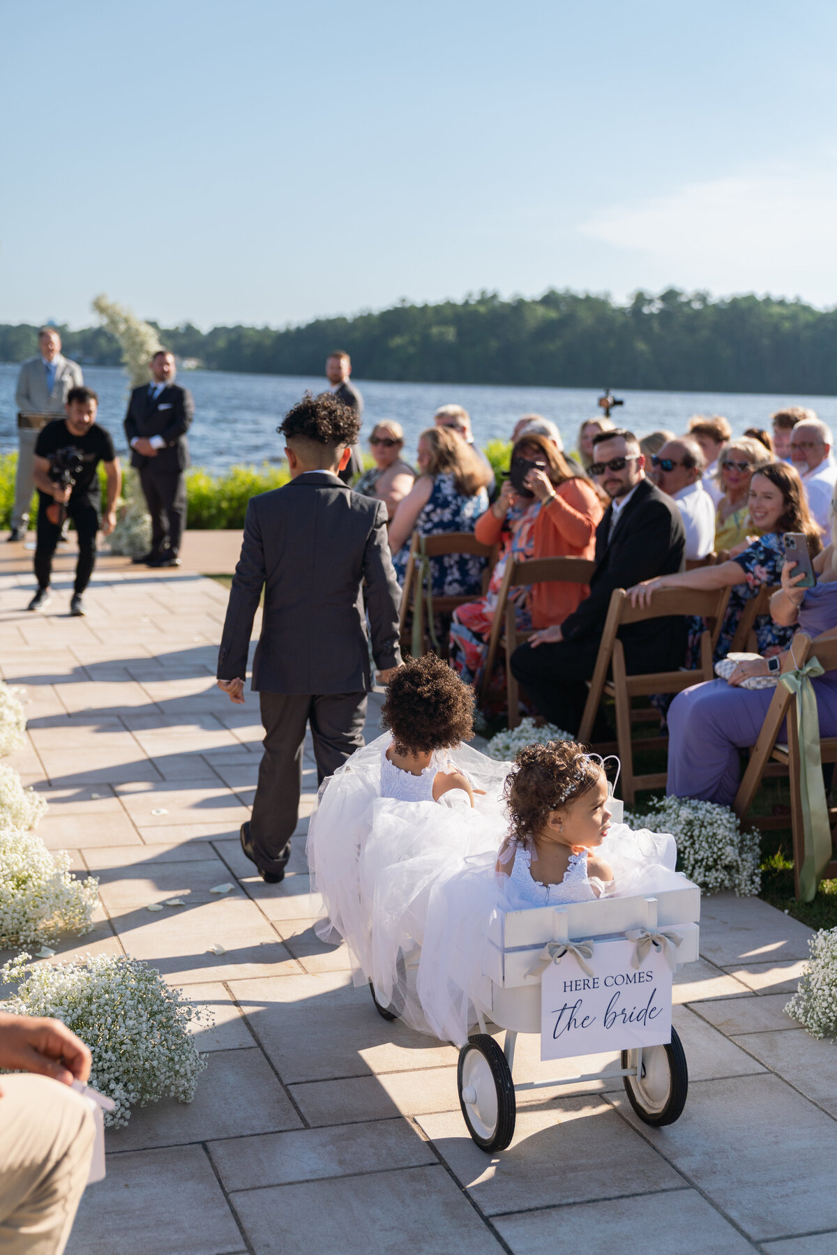 The Lakehouse Inn MA  | Kelsey Sheehan Photography Timeless Rhode Island Weddings | A young boy in a suit walks ahead of two girls in white dresses sitting in a wagon labeled "Here Comes the Bride." Guests seated outdoors by a lake watch them.