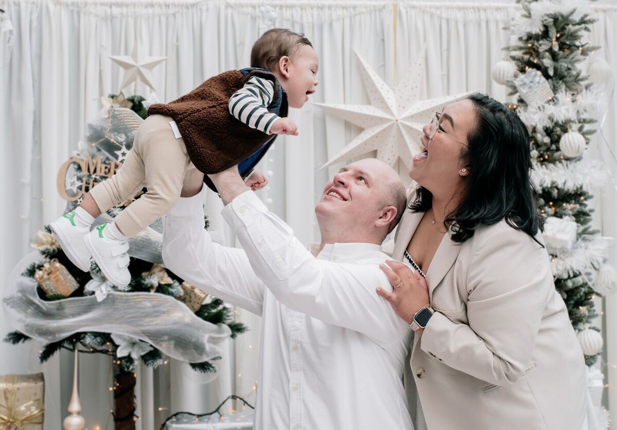 Parents lifting baby at Christmas – Joyful couple indoors by a decorated tree as the father lifts their smiling baby high in the air.