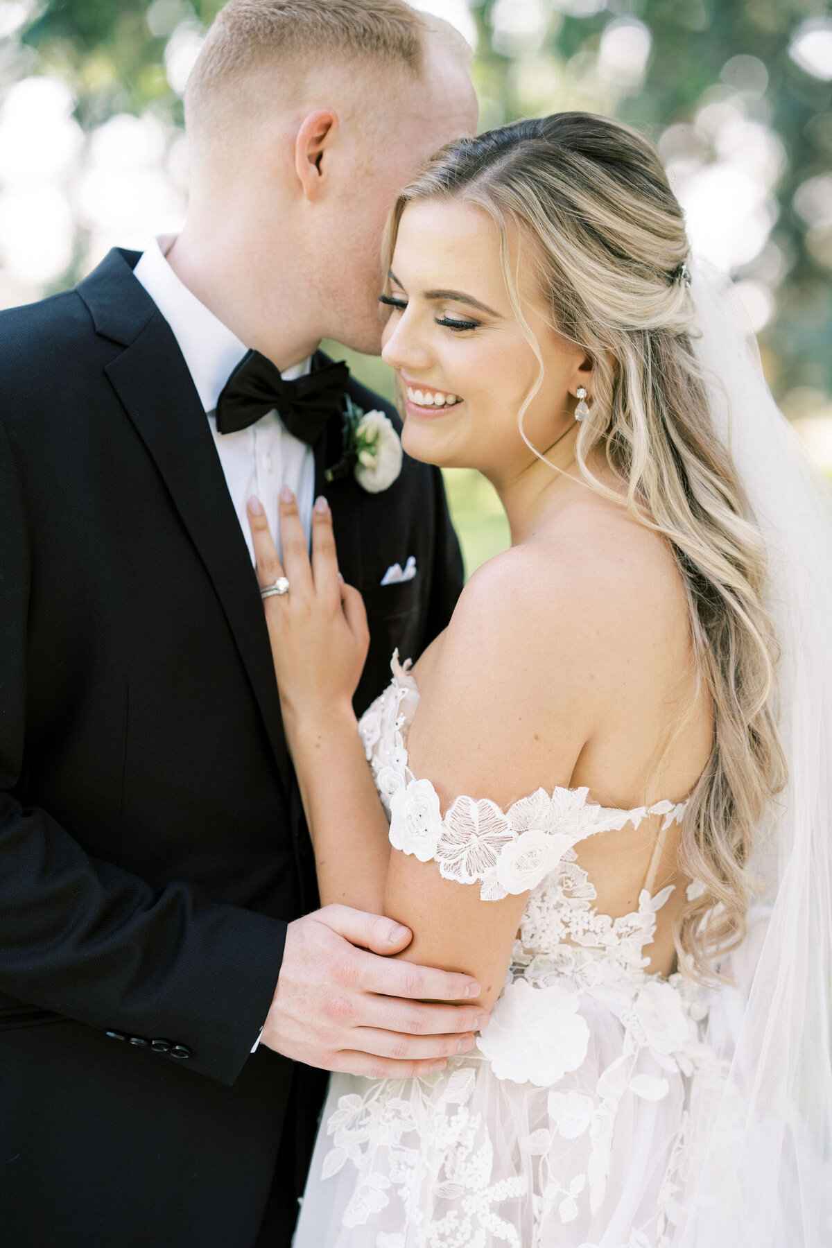 A bride and groom stand closely together outdoors at their wedding in Calgary at Norland Historic Estate. The bride smiles while the groom leans in. She wears a detailed white gown; he is in a dark suit with a bow tie and boutonniere.