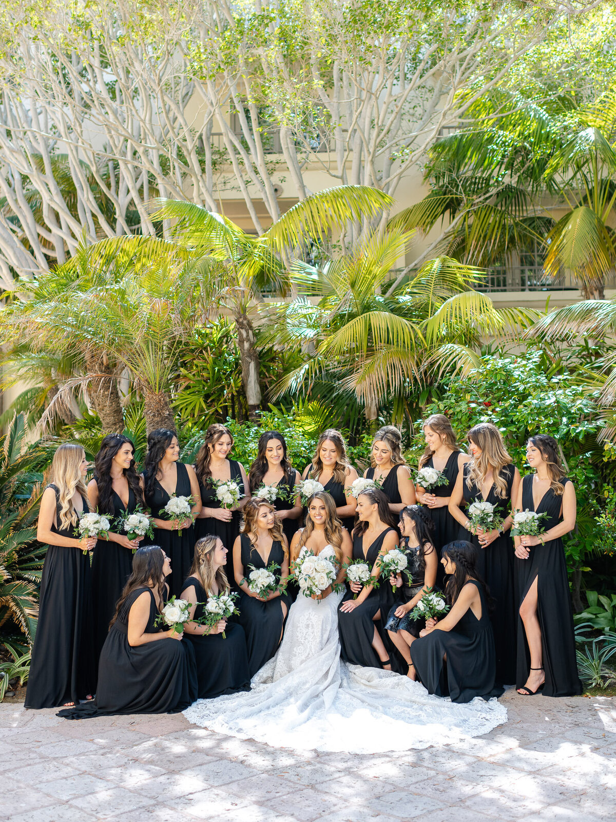 The bride, seated in her wedding gown, is surrounded by bridesmaids in black dresses, all holding white floral bouquets at Ritz-Carlton Laguna Niguel